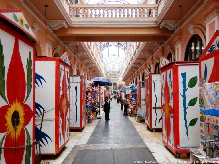 Market aisle beneath a glazed roof, with red-trimmed stalls painted with large flowers and shoppers receding into the passage.
