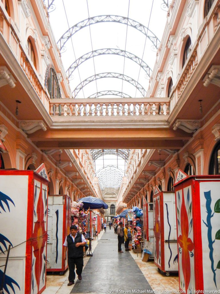 Covered market passage with peach arcades, glass-and-metal roof, painted flower stalls, vendors, and shoppers along a central aisle.