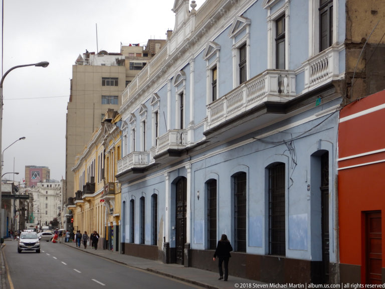 Oblique Lima streetscape with blue, yellow, and red historic façades, balconies, pedestrians, and vehicles.