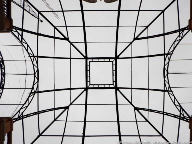 Upward view of dark geometric metal canopy framework with central rectangle, radiating members, and curved side sections.