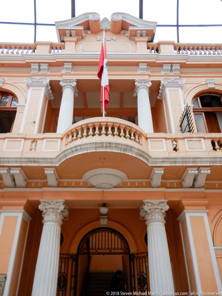 Upward view of peach façade with white columns, curved balcony, arched entrance, balustrades, and Peruvian flag.