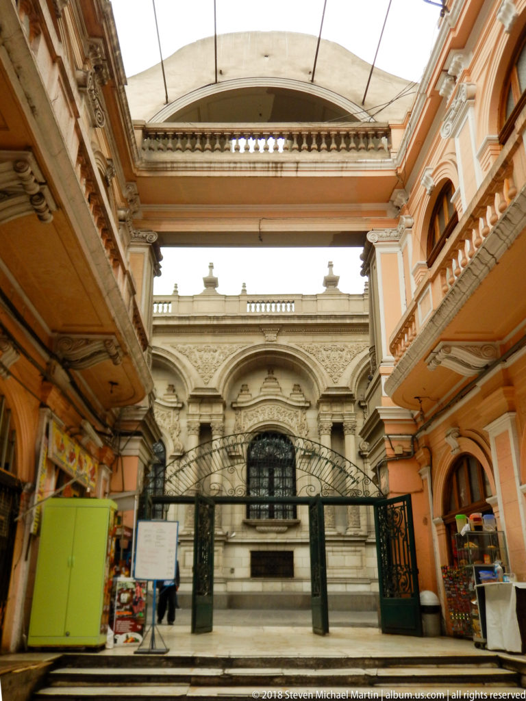 Peach-colored arcaded passage with shops leading through wrought-iron gates toward an ornate pale stone façade.