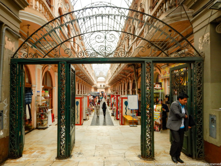 Decorative wrought-iron gate opening into a long arched shopping passage with stalls, displays, and pedestrians.
