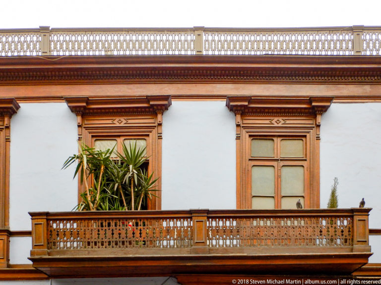 Long carved wooden balcony with patterned balustrade, tall wood-framed windows, plants, and white plaster wall.