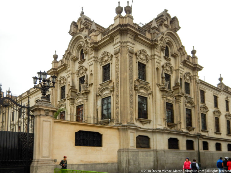 Ornate pale stone corner building with elaborate window surrounds, pilasters, cornices, perimeter wall, and street lamps.