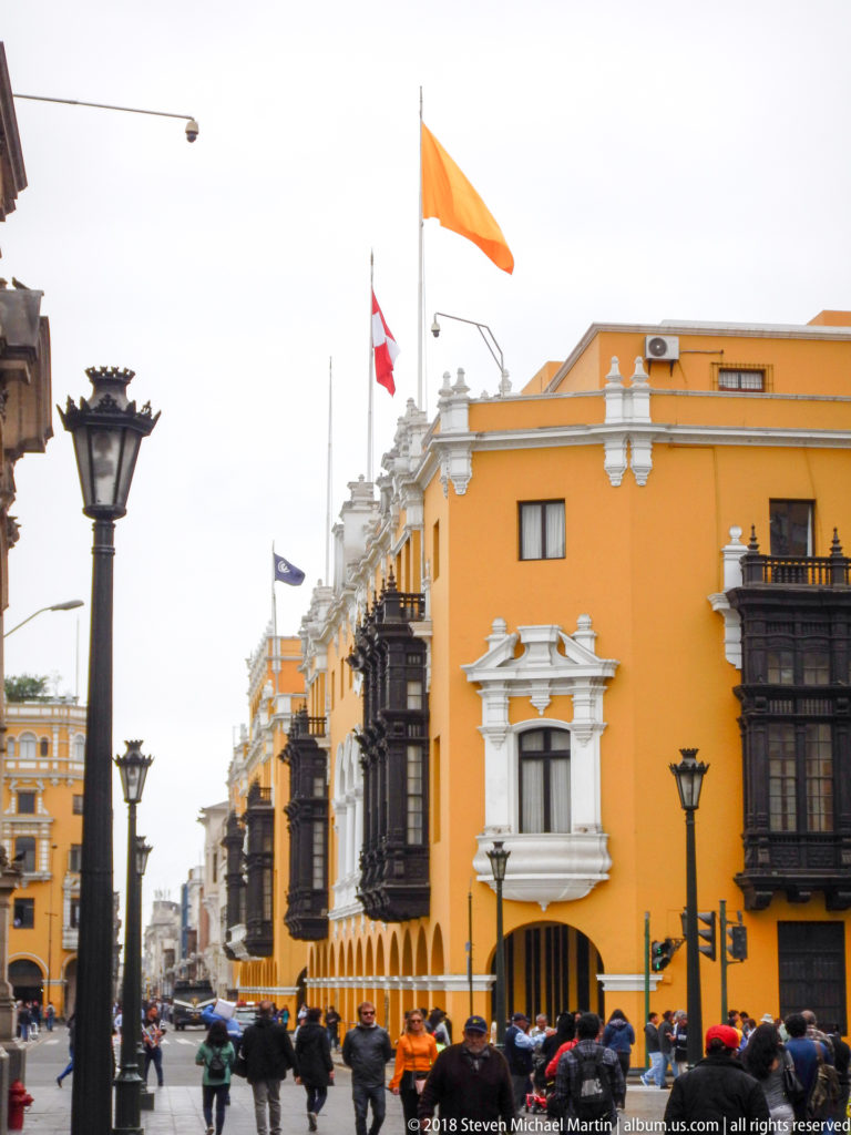 Ochre historic corner building with dark wooden balconies, white trim, flags, street lamps, cars, and pedestrians.