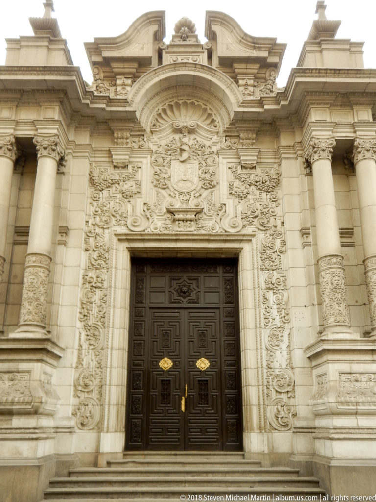 Dark double door framed by elaborate pale stone columns, relief carving, scrollwork, and layered architectural ornament.