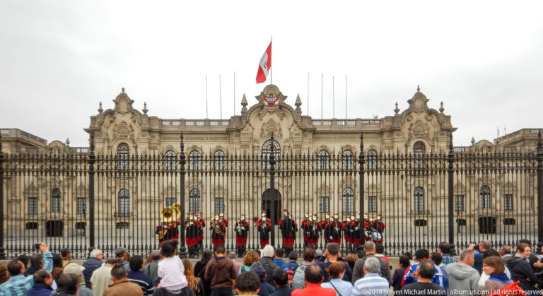 Crowd watching ranks of ceremonial guards behind the Government Palace fence, with palace façade and Peruvian flag behind.