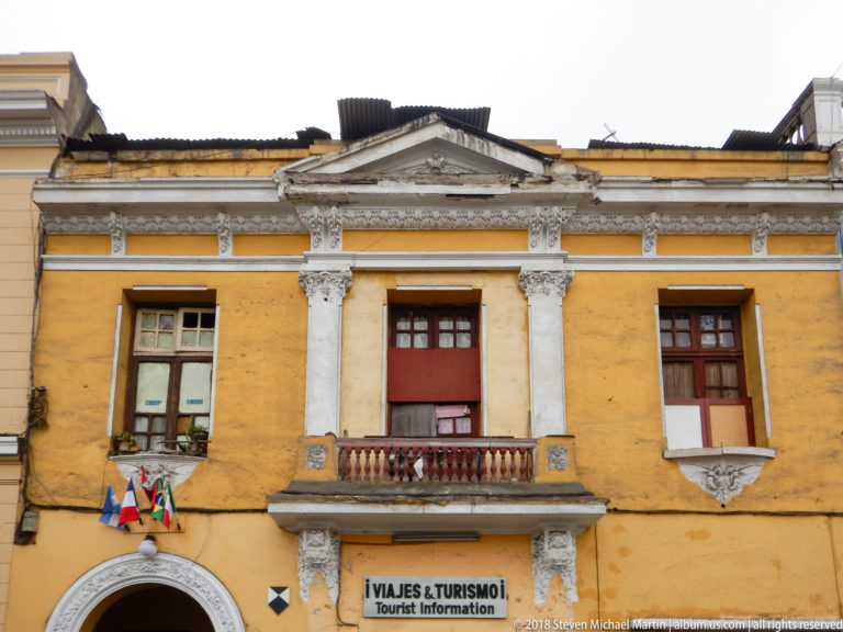 Ochre historic façade with ornate white trim, small balcony, flags, and a Viajes & Turismo Tourist Information sign.
