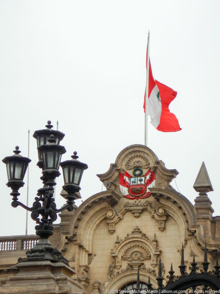 Peruvian flag flying above the national coat of arms and ornate stone crest, with a black street lamp in foreground.