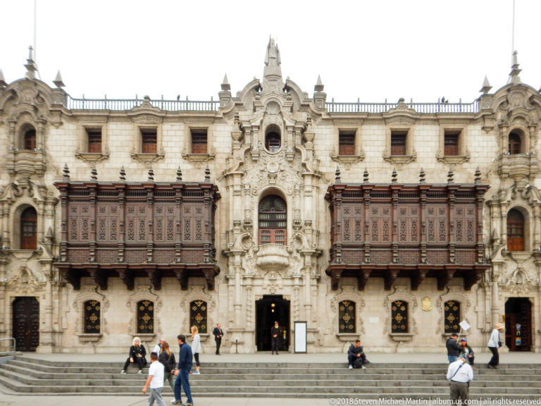 Ornate Archbishop’s Palace façade with two large carved wooden balconies, central stone portal, grilles, and pedestrians.
