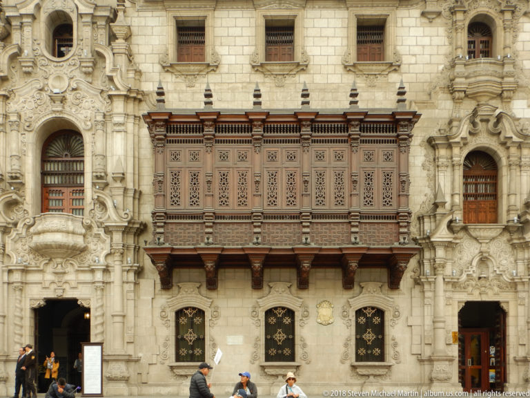 Large enclosed wooden balcony on ornate Archbishop’s Palace façade, with carved stonework, grilles, and pedestrians below.