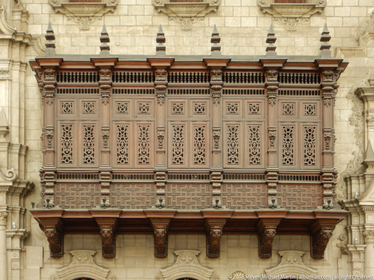 Long carved wooden enclosed balcony with lattice panels and timber brackets projecting from an ornate pale stone façade.