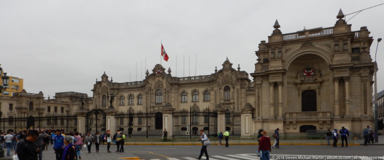Government Palace behind iron fencing with projecting monumental gateway and pedestrians gathered along the street.
