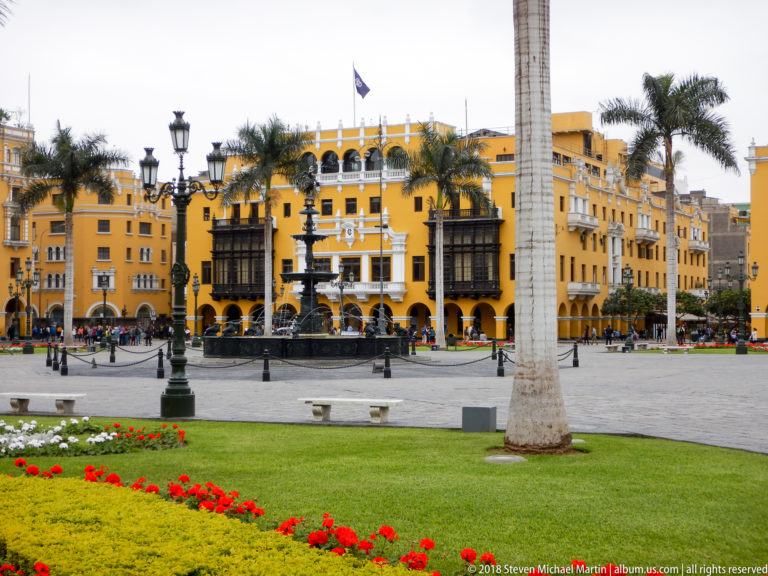 Red flower bed and lawn before Plaza Mayor fountain, palm trunk, and ochre building with dark balconies.