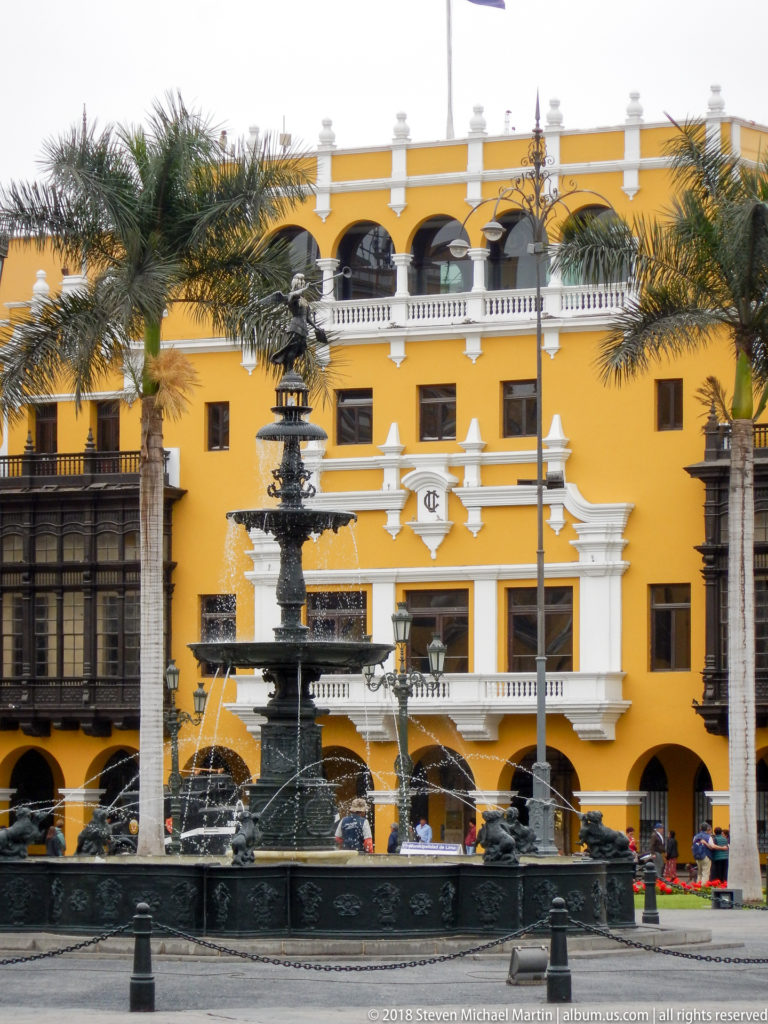 Dark multi-tiered fountain spraying water before an ochre building with white trim, balconies, arcades, and palms.