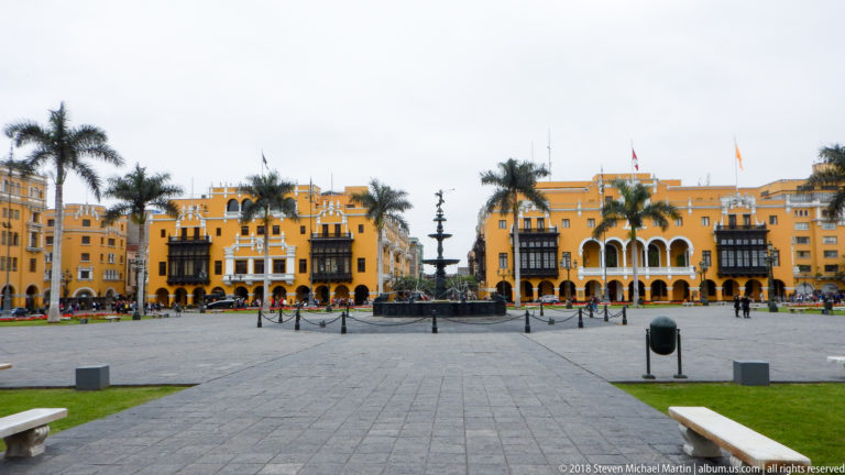 Centered paved walkway leading to Plaza Mayor fountain, with lawns, palms, and ochre balconied buildings beyond.