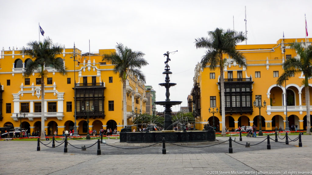 Bronze fountain in Plaza Mayor before ochre buildings with dark wooden balconies, arcades, palms, and flower beds.