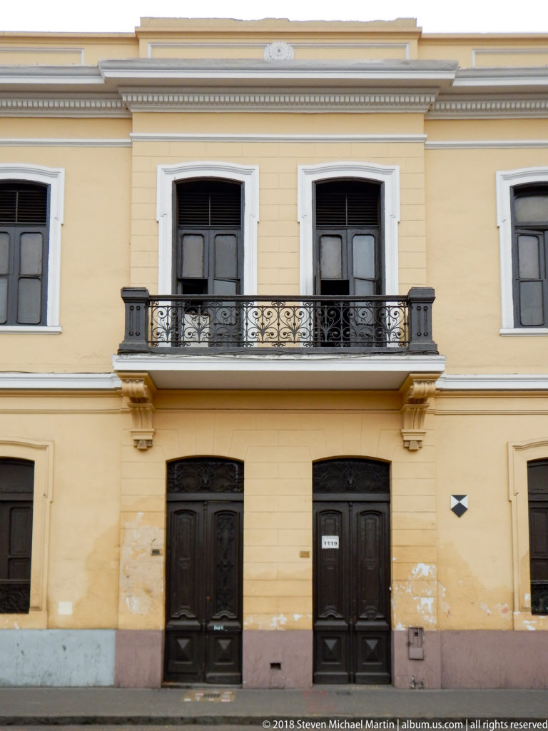 Yellow two-story historic façade with dark wooden doors and windows, white trim, and a central wrought-iron balcony.