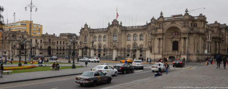 Oblique view of Government Palace beside Plaza Mayor with cars, pedestrians, iron fencing, and ceremonial entrance.