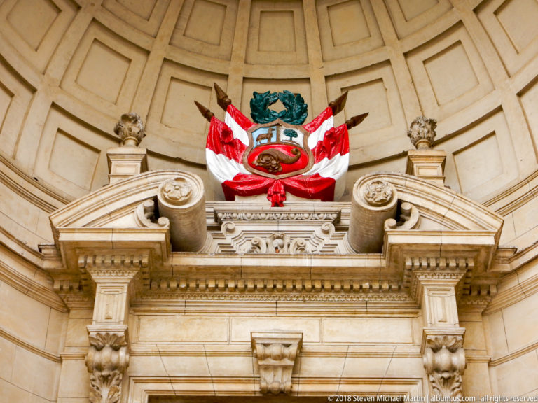 Peruvian coat of arms with red-and-white drapery above an ornate stone doorway framed by scrollwork.