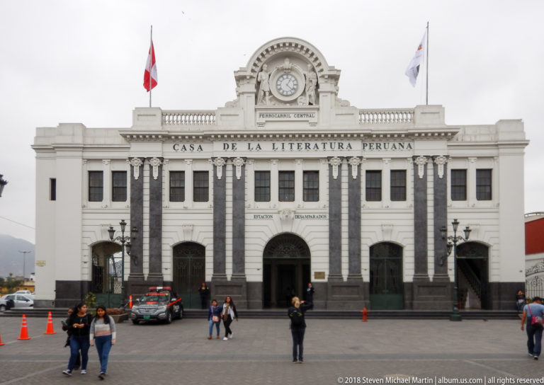 White façade of Casa de la Literatura Peruana with arched entrances, dark vertical accents, flags, and pedestrians.