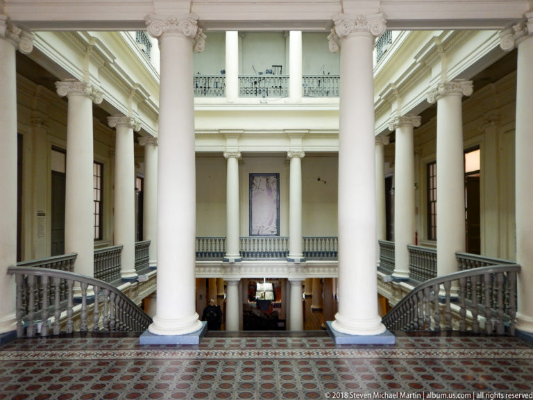 Two-story atrium with tall white columns, paired curved staircases, balustraded galleries, and patterned tile floor.