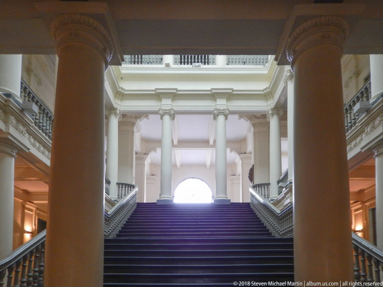 Broad interior staircase rising between large columns toward an arched opening, with symmetrical balustrades and upper galleries.
