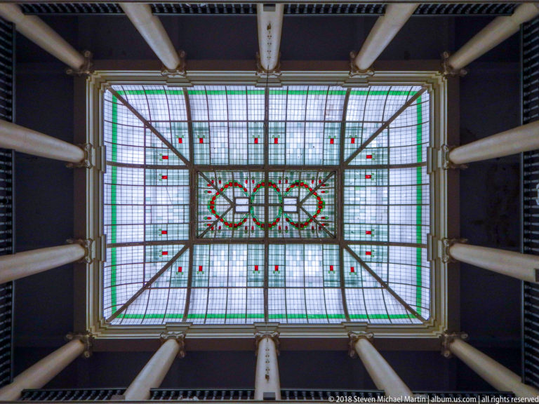 Rectangular decorative glass skylight viewed upward between pale columns, with geometric panes and circular motifs.