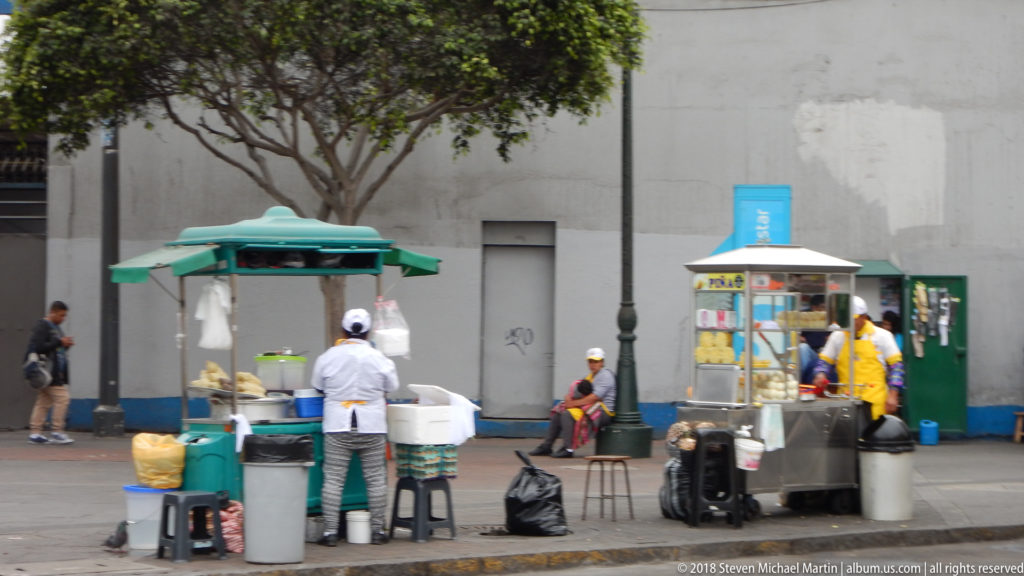 Two street-food carts with vendors, stools, bins, and a seated pedestrian along a broad Lima sidewalk.