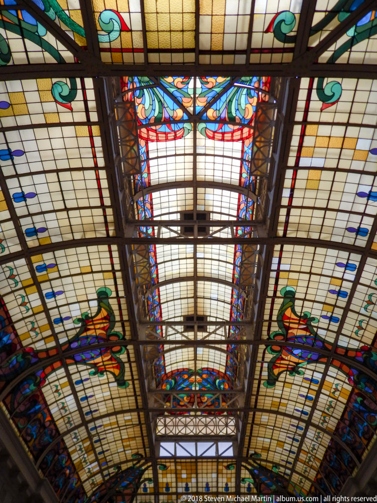 Axial close view of stained-glass canopy with dark ribs, pale rectangular panes, colorful floral borders, and a central roof opening.