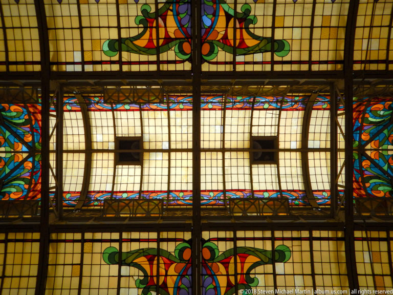 Close view of stained-glass ceiling with two large oval panels, cream rectangular panes, dark framing, and colorful floral borders.