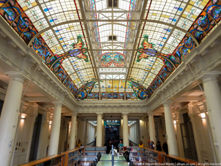 Spacious columned hall with visitors below a large colorful stained-glass roof and upper gallery.