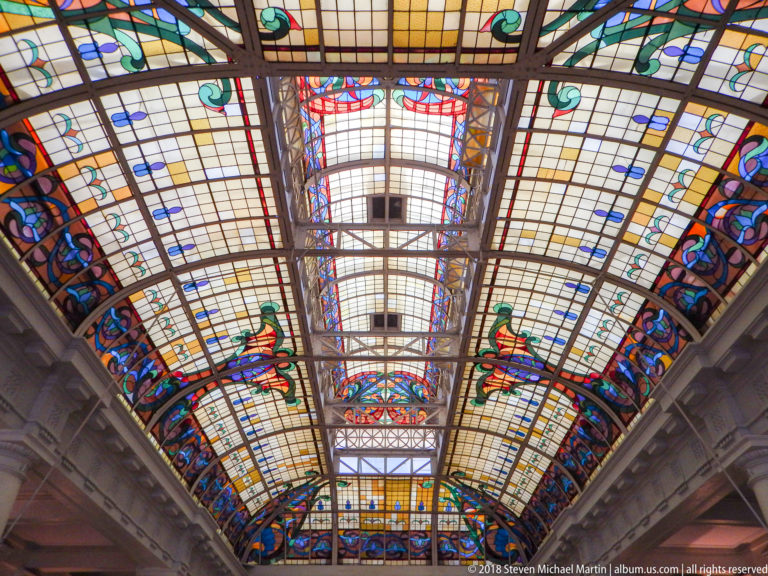 Long stained-glass ceiling with cream grid panels, colorful floral borders, and dark structural ribs viewed from directly below.