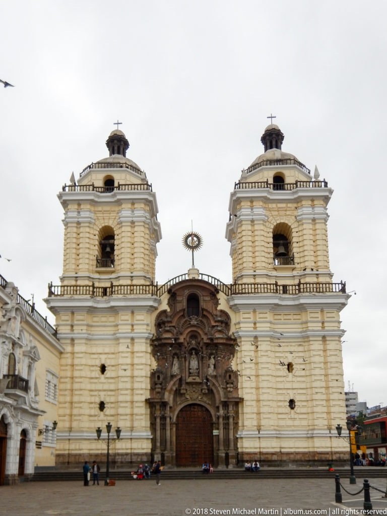 Frontal view of a pale twin-towered colonial church with an ornate dark central portal and open plaza in front.