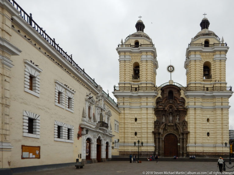 Cream colonial church with twin bell towers and a dark ornate central portal facing an open paved plaza.