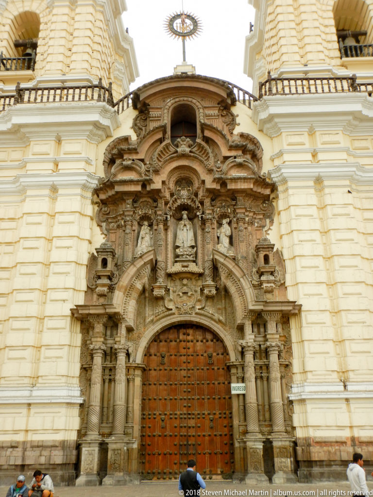 Elaborately carved baroque stone church portal with niches, sculpted figures, layered columns, and a large studded wooden door.