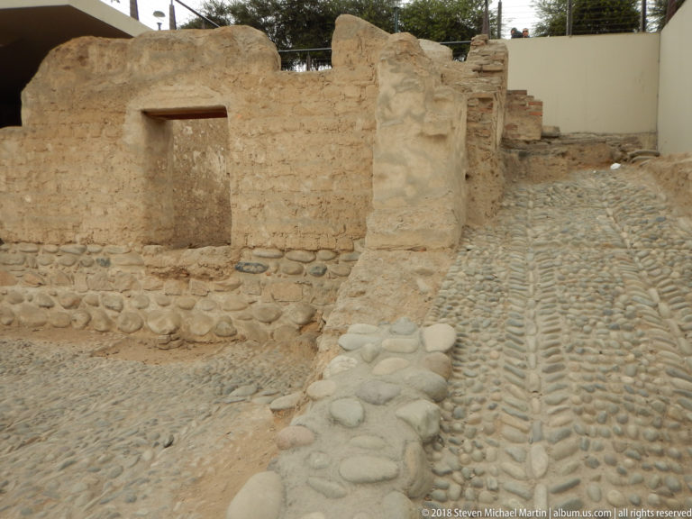 Low eroded adobe walls and cobble paving or foundations within an archaeological area in Lima.