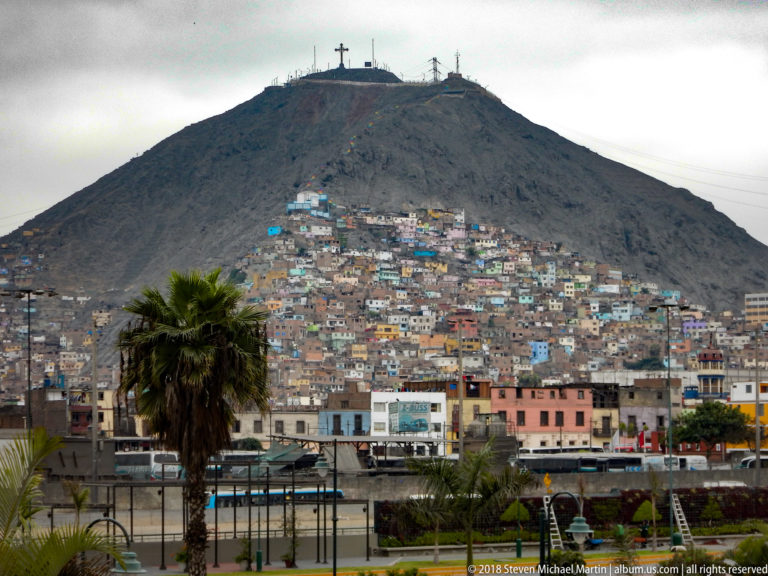 Cerro San Cristóbal rising above dense hillside housing, with a large cross on the barren summit and palms in the foreground.