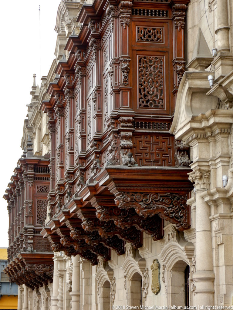 Oblique view along several dark carved wooden balconies projecting from the pale stone façade of the Archbishop’s Palace.