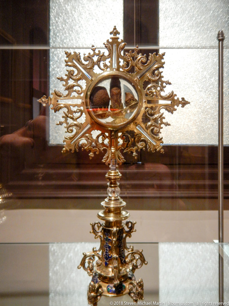 Gilt reliquary with oval glazed center, radiating openwork frame, ornate stem, and colored decorative accents.