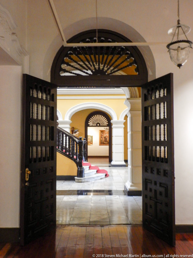 Open fanlight doors frame white arches, yellow walls, and a distant red-carpeted staircase across the palace hall.