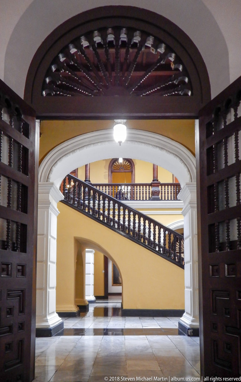 Open dark doors frame a sweeping staircase with yellow wall, white arches, and dark wooden balustrade.