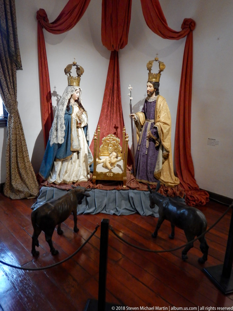 Two crowned religious statues in ornate robes stand beneath red drapery with dark animal sculptures in front.