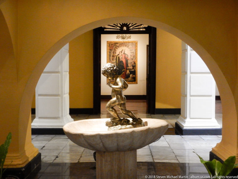 Shallow stone basin beneath an arch supports a kneeling bronze figure holding flowers, with framed art beyond.