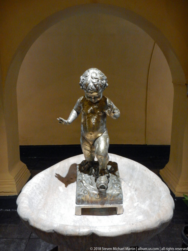 Bronze child figure with outstretched arms on an ornamented pedestal inside an arched niche.