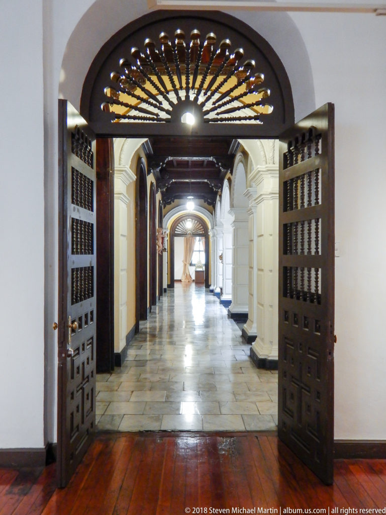 Open dark doors and radial fanlight frame a long stone-floored corridor lined with white arches and dark doorways.