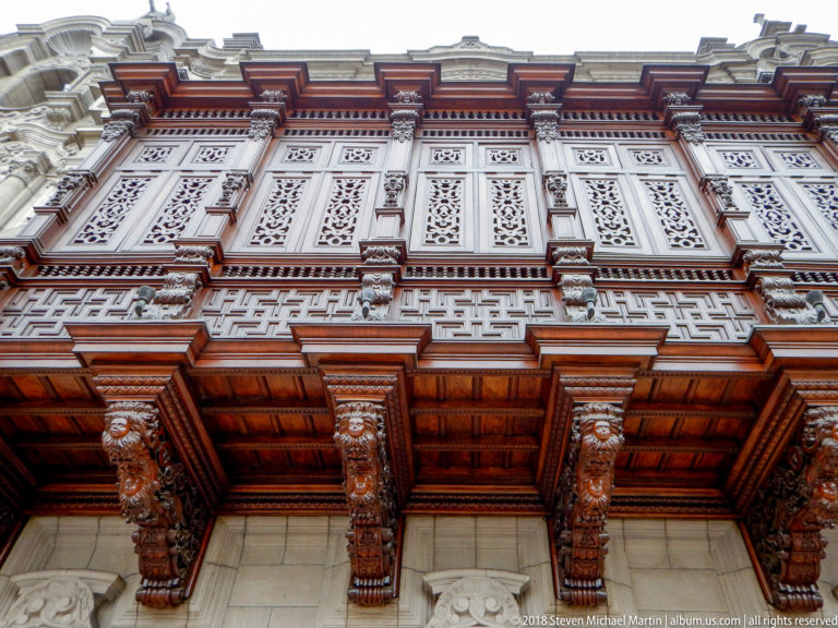 Upward view of an ornate projecting wooden balcony with carved brackets, geometric panels, latticework, and pale stone façade.