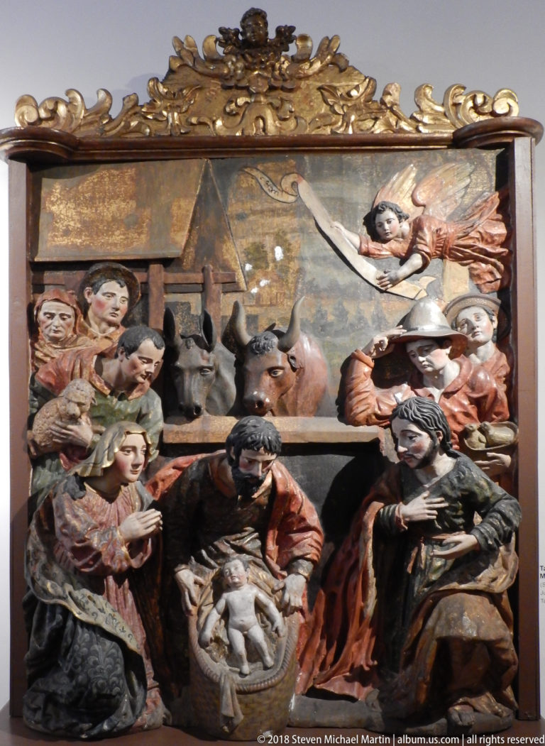 Polychrome Nativity relief with infant, Mary and Joseph, ox, donkey, attendants, and an angel above.