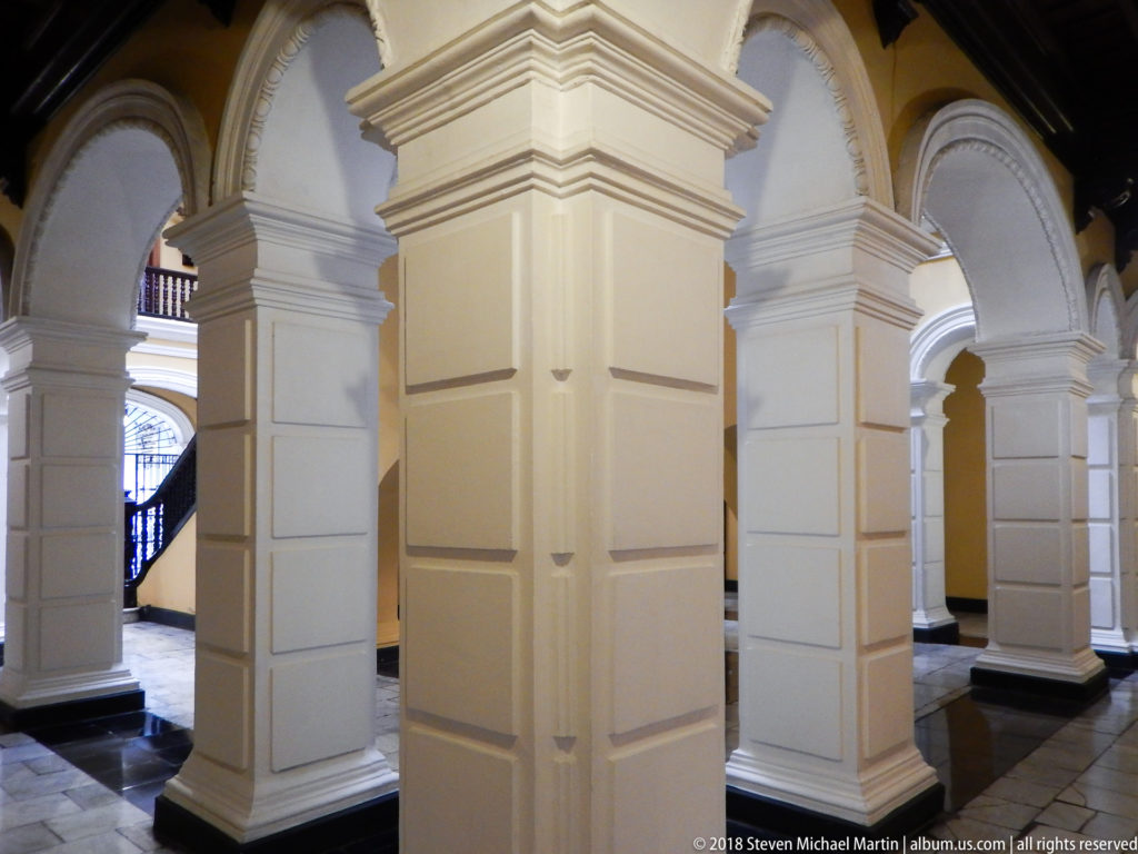 White square piers and rounded arches line a stone-floored palace corridor with glimpses of stairs beyond.